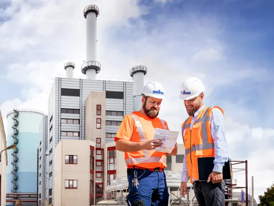 Zwei Männer mit Warnweste und Helm stehen vor dem Heizkraftwerk Schwerin Süd und schauen auf einen Plan.