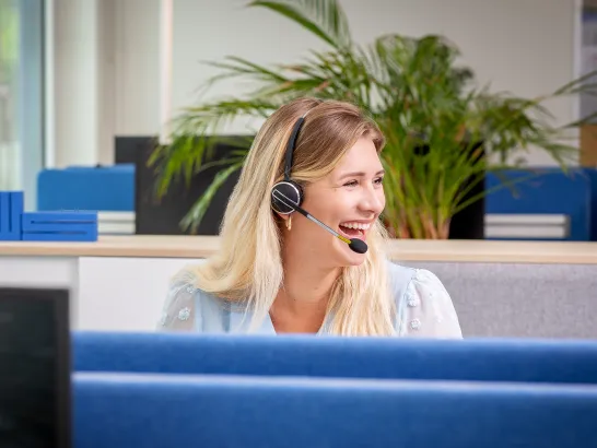 Eine junge Frau mit langen blonden Haaren und Headset sitzt am Schreibtisch.