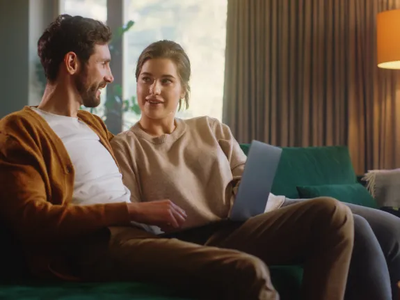 Junges Pärchen sitzt mit dem Laptop auf dem Sofa und schaut sich an.
