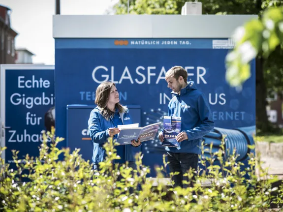 Zwei Stadtwerke-Mitarbeiter unterhalten sich mit Unterlagen in der Hand vor einem Glasfaser-Multifunktionsgehäuse.
