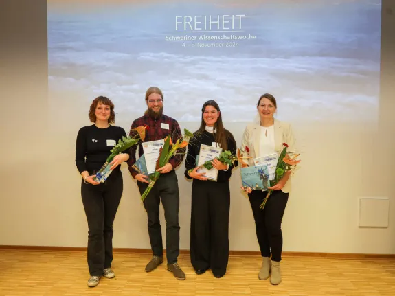 Preisträger stehen mit Blumen und Urkunden vor einer Leinwand.