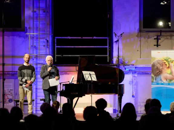 Zwei Herren stehen am linken Rand einer bunt ausgeleuchteten Bühne mit Piano.
