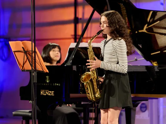 Junge Musikerin mit Saxophon wird im Hintergrund von ihrer Lehrerin am Klavier begleitet