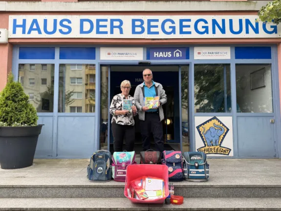 Ein Mann und eine Frau stehen mit gespendeten Ranzen vor einem Haus