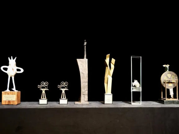 Sieben Trophäen der unterschiedlichen Kategorien des Filmkunstfestes MV stehen aufgereiht vor einem schwarzen Hintergrund.