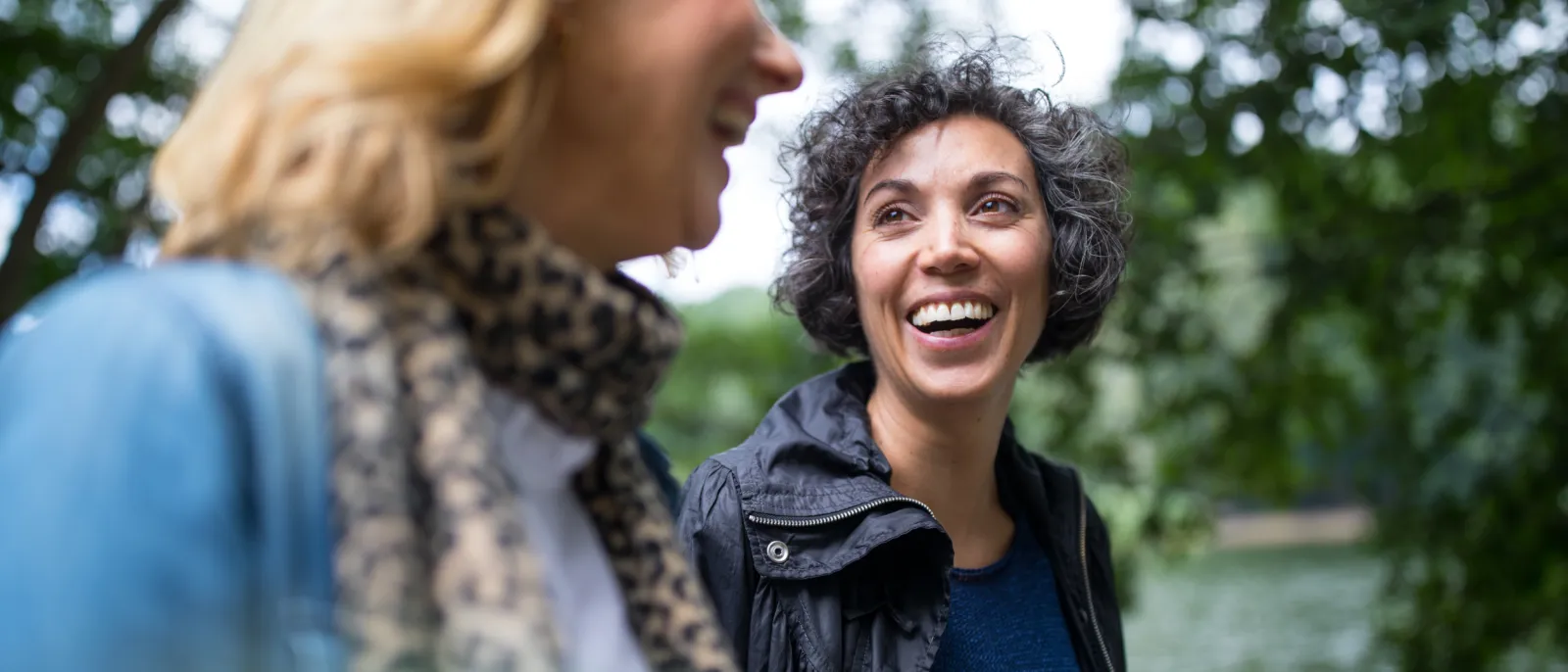 Zwei Freundinnen gehen im Wald spazieren und lächeln sich an.