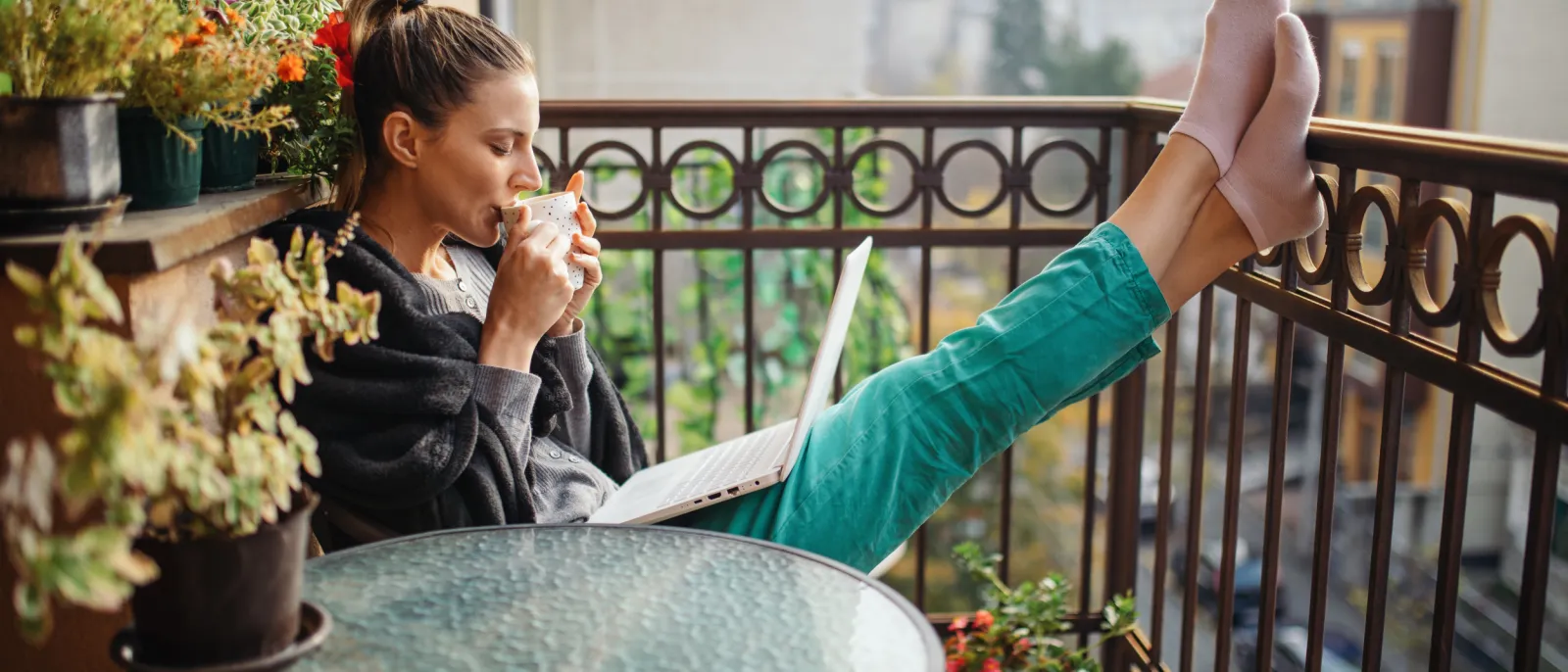Eine junge Frau sitzt mit ihrem Laptop auf dem Balkon und genießt eine Tasse Kaffee.