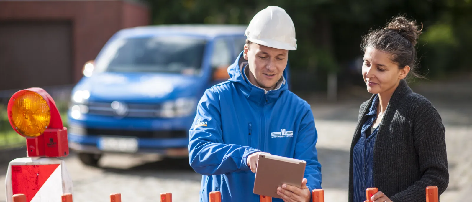 Ein Stadtwerke-Mitarbeiter zeigt einer Frau Informationen auf einem Tablet-PC. Sie stehen an einer Baustelle.