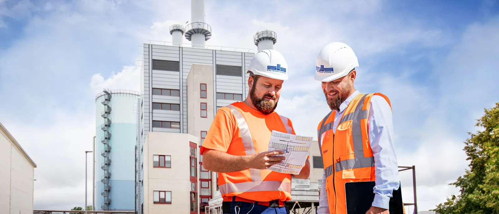 Zwei Mitarbeiter mit Helm und Warnweste stehen vor dem Heizkraftwerk und schauen gemeinsam auf einem Plan.