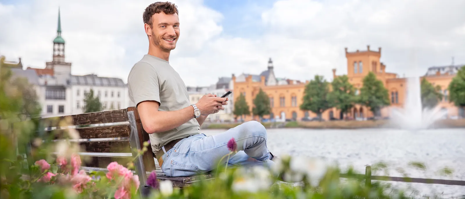 Ein Mann sitzt auf einer Bank vor dem Schweriner Pfaffenteich und surft auf dem Handy.