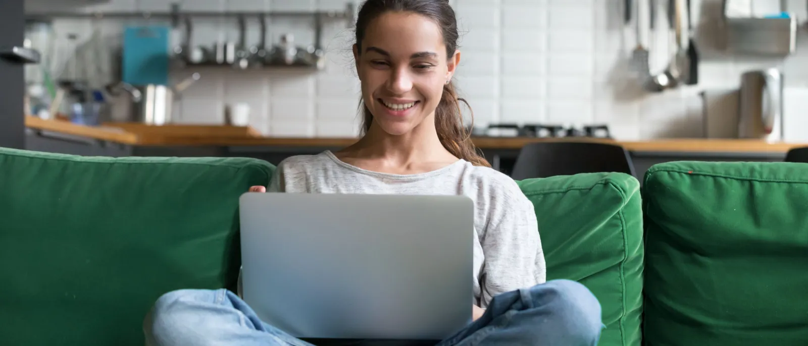 Eine Frau sitzt lächelnd im Schneidersitz auf dem Sofa mit einem Laptop auf dem Schoß.