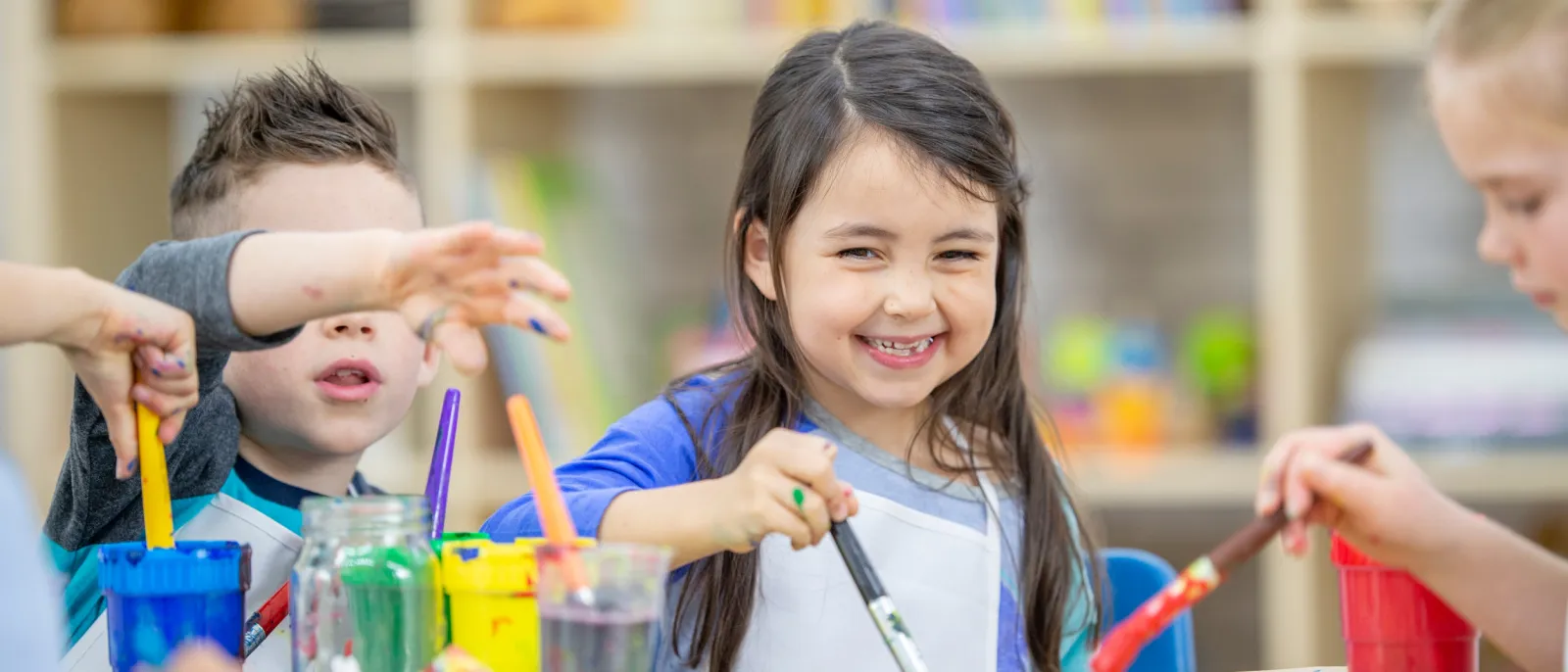 Nahaufnahme von drei Kindern im Vorschulalter beim gemeinsamen Malen mit verschiedenen Farben, Mädchen mit langen schwarzen Haaren steht im Mittelpunkt und schaut in die Kamera