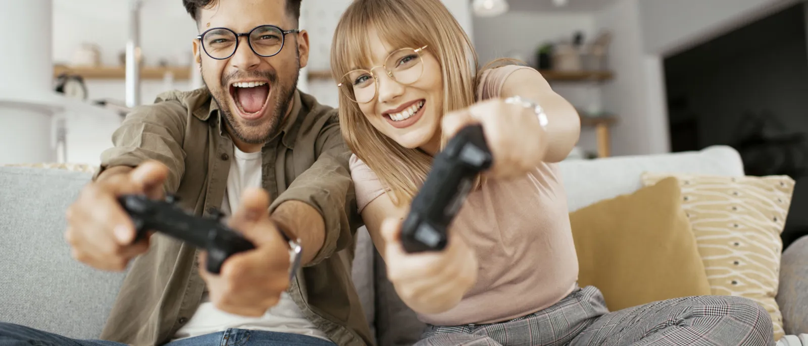 Ein Mann und eine Frau sitzen mit Controllern auf der Couch und gucken fröhlich in die Kamera.