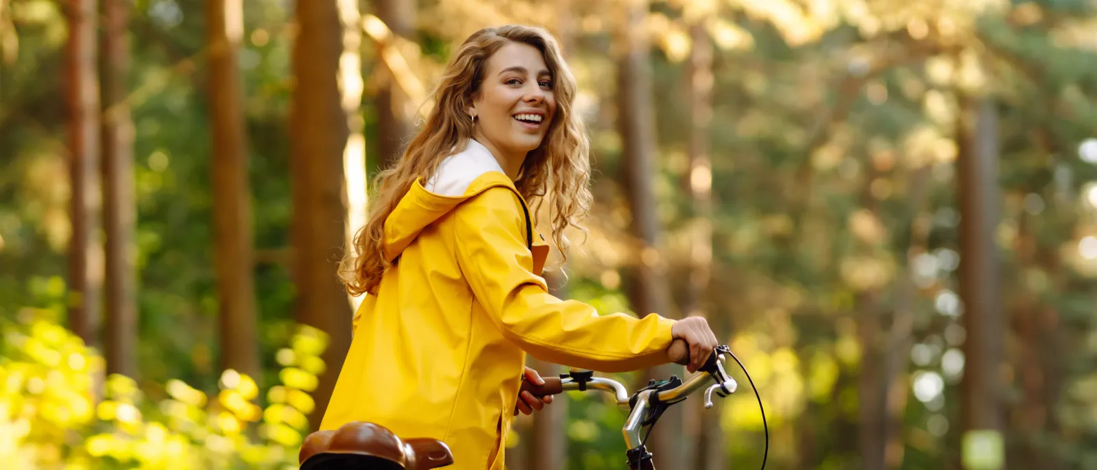Eine Frau im gelben Mantel fährt mit Ihrem Fahrrad durch einen Wald.