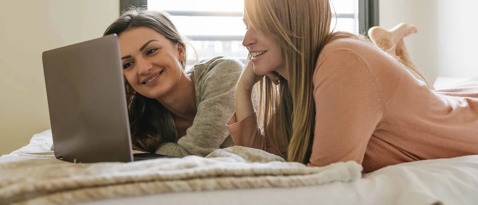Zwei junge Frauen liegen auf dem Bett und sehen auf einen Laptop.