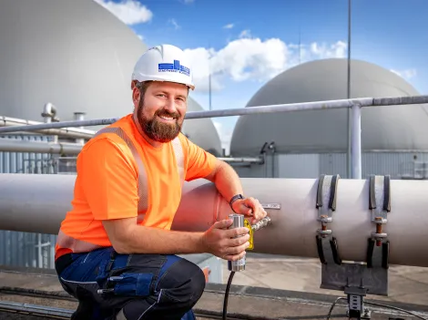 Ein Mitarbeiter in oranger Arbeitskleidung kniet auf dem Dach einer Biogasanlage.