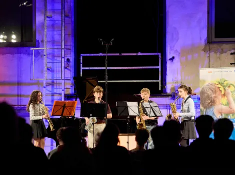 Vier junge Musiktalente mit Blasinstrumenten stehen auf einer blau beleuchteten Bühne