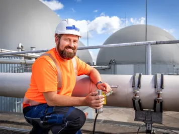 Ein Mitarbeiter in oranger Arbeitskleidung kniet auf dem Dach einer Biogasanlage.