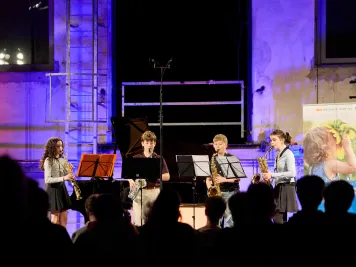 Vier junge Musiktalente mit Blasinstrumenten stehen auf einer blau beleuchteten Bühne