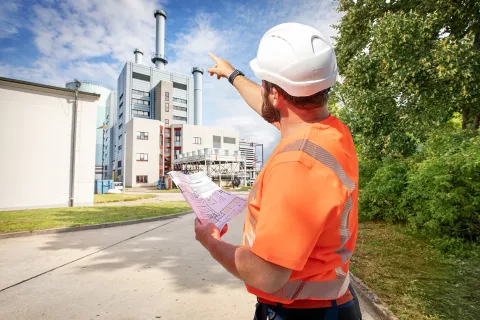 Ein Mann mit Warnweste und Helm zeigt mit dem Finger auf das Heizkraftwerk Schwerin Süd.