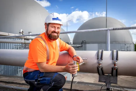 Ein Mann mit Warnweste und Helm kniet vor einer großen Rohrleitung vor der Biogasanlage in Schwerin Süd.