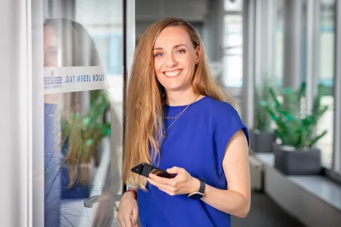 Eine junge Frau steht lächelnd in einer offenen Tür. Sie hält ein Smartphone in der Hand.