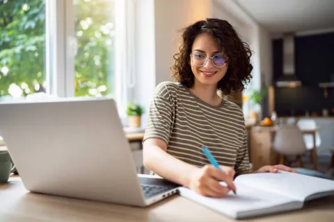 Junge Frau sitzt vor ihrem Laptop am Schreibtisch. Sie schreibt etwas auf einen Zettel.