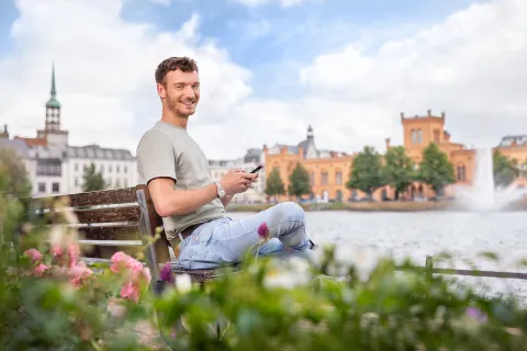 Ein Mann sitzt auf einer Bank vor dem Schweriner Pfaffenteich und surft auf dem Handy.