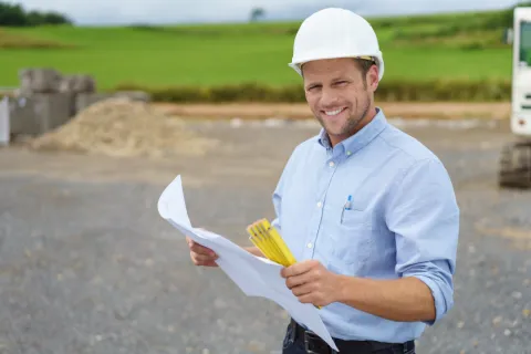 Ein Bauarbeiter mit einem Bauplan in der Hand lächelt in die Kamera.
