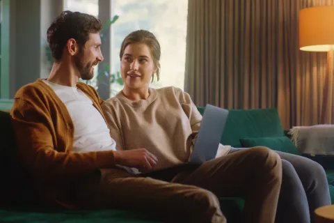 Ein junges Pärchen sitzt mit dem Laptop auf dem Sofa und schaut sich an.