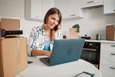 Eine Frau sitzt zwischen Umzugskartons vor ihrem Laptop in der Küche.