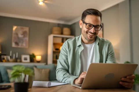 Ein Mann sitzt im Wohnzimmer vor dem Laptop und lächelt.