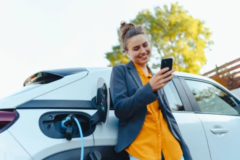 Junge Frau lehnt mit dem Handy in der Hand am Auto, während dieses lädt.