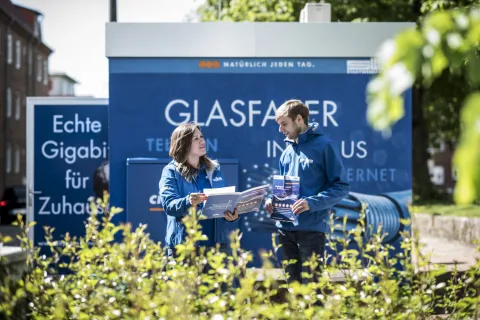 Zwei Stadtwerke-Mitarbeiter unterhalten sich mit Unterlagen in der Hand vor einem Glasfaser-Multifunktionsgehäuse.