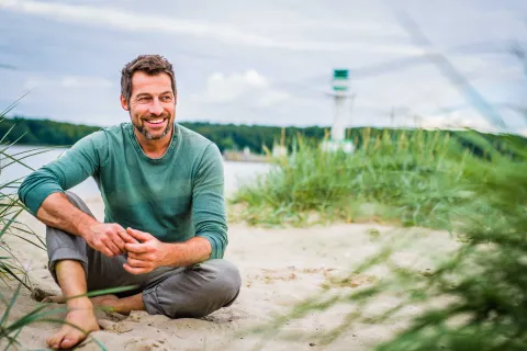 Ein Mann sitzt entspannt am Strand. Hinter ihm steht ein Leuchtturm.