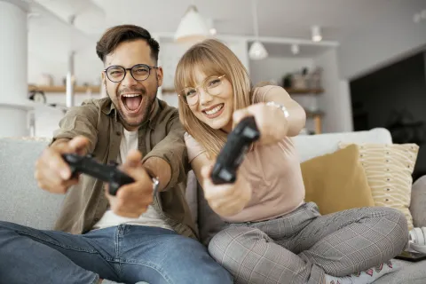 Ein Mann und eine Frau sitzen mit Controllern auf der Couch und gucken fröhlich in die Kamera.