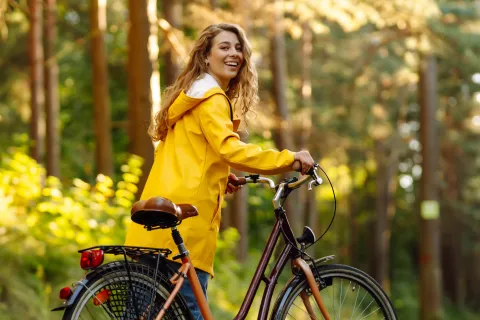 Eine Frau im gelben Mantel fährt mit Ihrem Fahrrad durch einen Wald.