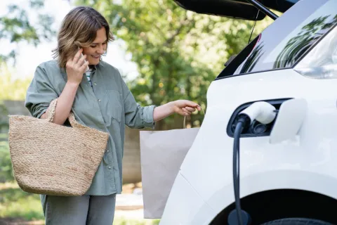 Frau steht mit Einkaufstasche neben ihrem E-Auto, das gerade lädt