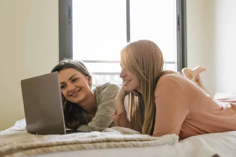 Zwei junge Frauen liegen auf dem Bett und sehen auf einen Laptop.