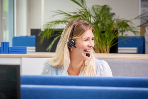 Eine junge Frau mit langen blonden Haaren und Headset sitzt am Schreibtisch.