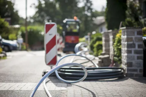 Leerrohre für Glasfaser liegen neben einer Baustellenbake auf einem Gehweg mit Minibagger im Hintergrund.
