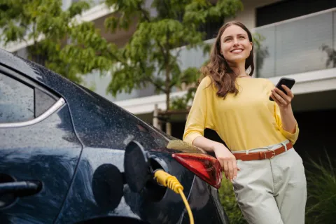 Frau steht mit Handy in der Hand neben einem ladenden E-Auto