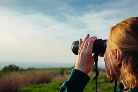 Eine Wanderin schaut mit dem Fernglas auf eine Küstenlandschaft.