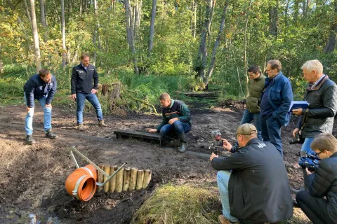 Acht Männer gruppieren sich um ein Stauwerk im Wald, in der Mitte kniet ein Landesforst Mitarbeiter und erklärt.