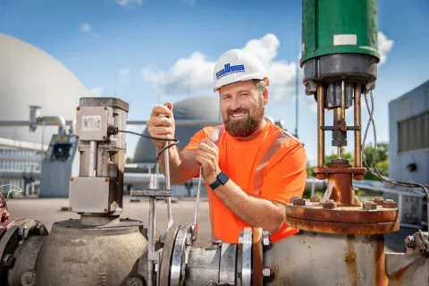 Mitarbeiter mit weißem Helm und Stadtwerkelogo vor den Kuppeln der Biogasanlage.