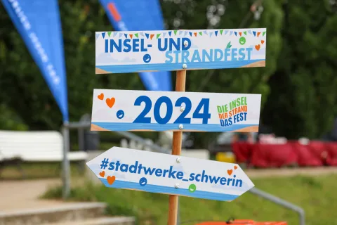 Wegweiser mit drei Hinweisschildern "Insel- und Strandfest", "2024" und "#stadtwerke_schwerin"