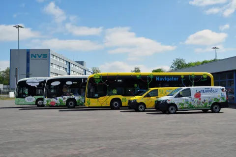 Drei E-Linienbusse mit unterschiedlichen Designs sowie zwei elektrische Transporter stehen vor dem Hauptgebäude des NVS.
