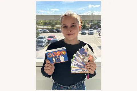Eine junge Frau hält lächelnd mehrere Postkarten aufgefächert in der Hand und präsentiert eine einzelne Karte.