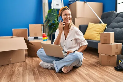 Junge Frau mit Laptop und Handy zwischen Umzugskartons.