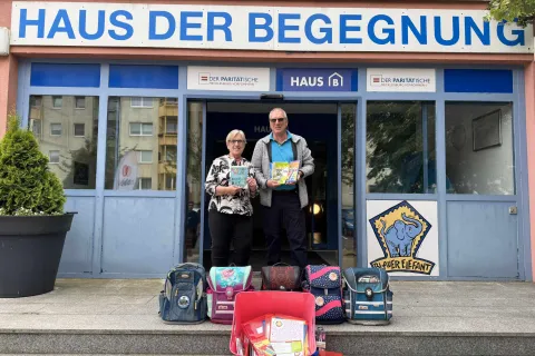 Zwei Personen stehen vor einem Haus und halten Schreibhefte in den Händen. Davor stehen Schulranzen und andere Schulmaterialien.
